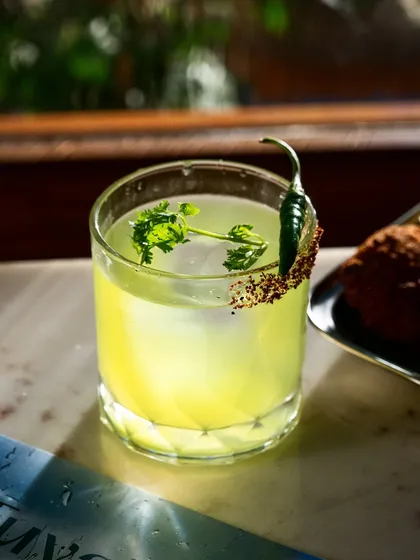 A spicy cilantro cocktail shot in natural light. The rim seasoning and fresh garnish are the heroes here, signaling a fresh and zesty flavor profile.