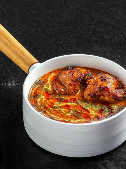 A hearty chicken curry in a white serving dish. The focus is on the thick, textured gravy and tender pieces of chicken.