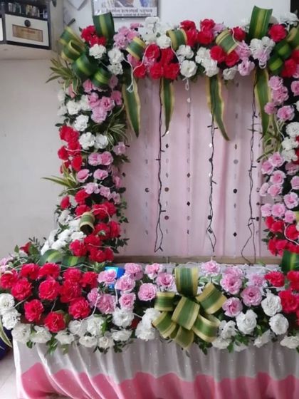 This dense floral frame is made with red, pink, and white roses and accented with green leaf-style ribbons for a traditional touch. The string lights woven into the pink backdrop add a subtle sparkle to the decoration.