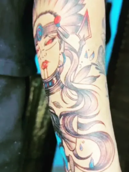 A forearm tattoo for girls, featuring a stylized portrait with a headdress. The touches of red and blue color add highlights to this illustrative black and grey piece.