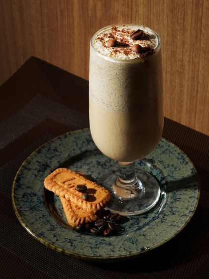 A tall glass of a coffee milkshake, styled with cookies and coffee beans. The dark, woody background gives it a cozy, cafe-like atmosphere.