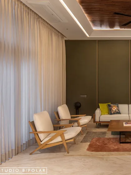 Sheer curtains diffuse natural light, creating a soft and airy feel that balances the deep green of the walls. The clean lines of the armchairs add to the modern aesthetic of the lounge.
