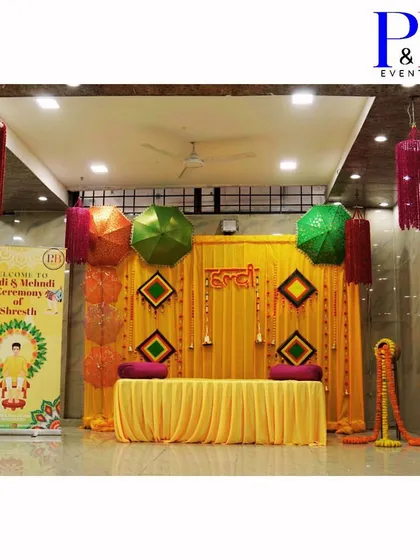 A complete Haldi and Mehendi setup in a banquet hall. The stage features a bright yellow backdrop with green umbrellas and traditional hangings, along with a personalized welcome standee for the groom.