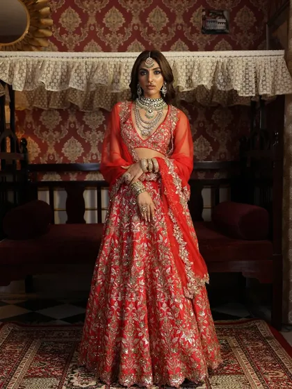 A full-length portrait against a vintage backdrop, enhancing the timeless quality of the red lehenga. The styling is designed to create a picture of classic elegance.