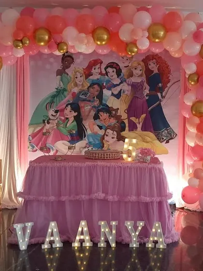 A Disney Princess theme featuring a large backdrop with multiple princesses. The decoration is completed with a pink and gold balloon arch and marquee letters.