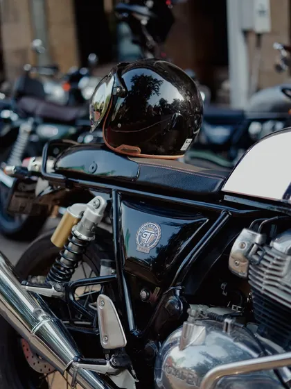 A glossy black helmet rests on the seat of a Royal Enfield Continental GT. This shot focuses on the details of the bike's tank, badge, and the lifestyle associated with cafe racer culture.
