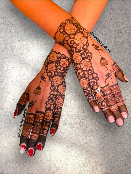 A stunning back-of-hand design that combines a dense floral vine with delicate hanging jhumkas. The contrast between the full pattern and the simple finger details is very effective.
