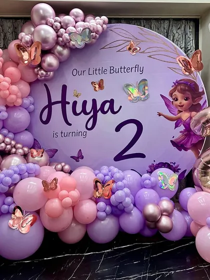 A magical butterfly-themed second birthday for "Hiya," featuring a large circular backdrop in soft purple. The decor is brought to life with a stunning organic balloon garland in shades of pink and purple, accented with metallic and iridescent butterfly cutouts and a large rose gold '2' foil balloon.
