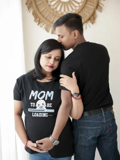 A sweet and tender moment between the couple in their matching "Mom to be" and "Dad to be" shirts. The natural light from the window creates a soft, gentle mood.