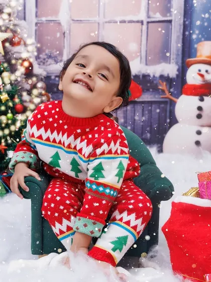 A cheerful toddler in a Christmas sweater sits on a miniature chair, surrounded by snow and presents.