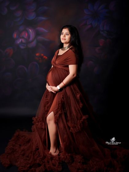 A classic solo maternity pose in a flowing brown gown. The side profile and soft lighting beautifully highlight the baby bump against a dark floral background.