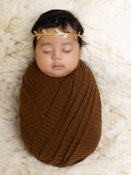 Wrapped in a deep brown swaddle, this sleeping newborn wears a golden leaf crown. The simplicity of the pose on a soft white fur rug makes for an elegant and classic portrait.