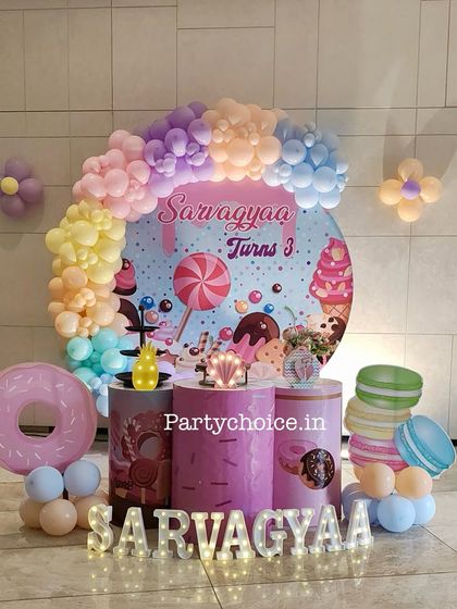 A sweet Candyland theme for a 3rd birthday. The setup is filled with colorful props like donuts, macarons, and lollipops, set against a backdrop of dripping ice cream and a pastel balloon arch.