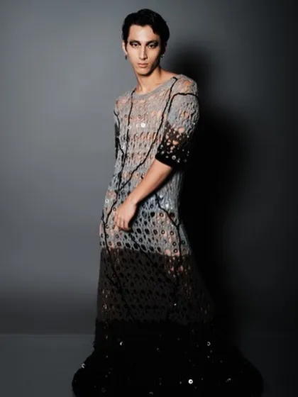 A full-length view of the men's editorial look. The sleek hair and bold makeup provide a sharp contrast to the soft, netted fabric of the outfit, creating a memorable fashion image.