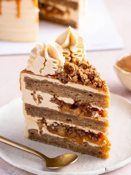 A slice of our Apple and Salted Caramel Cake, a true hug in cake form. You can see the layers of spiced apple sponge, gooey apple pie filling, and silky salted caramel buttercream, all topped with a crunchy crumble.