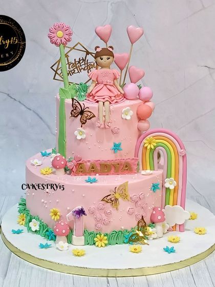 A charming two-tier choco-chip cake for a first birthday. This pink cake features a fondant doll, a rainbow, butterflies, and heart-shaped balloon accents for a sweet and lovely design.