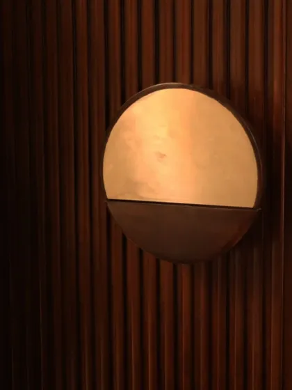 A detail of a custom wall sconce, where a simple circular brass plate is set against a dark, fluted wooden panel. This interplay of light, material, and texture creates a subtle yet sophisticated design moment.