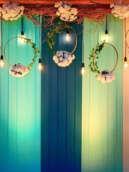 A close-up of a rustic-chic backdrop. The design combines a multi-toned wood panel with hanging floral wreaths and exposed bulbs, creating a perfect blend of natural and industrial styles for a Mehendi event.