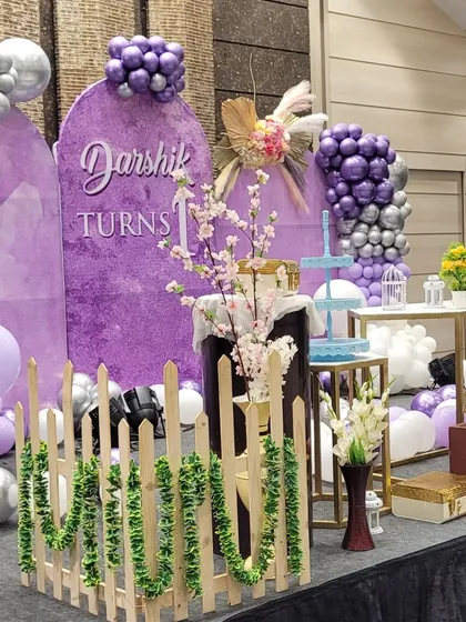 A side view of the purple-themed first birthday stage, showing the layered decor with a small picket fence, floral arrangements, and pedestals for the cake and desserts.