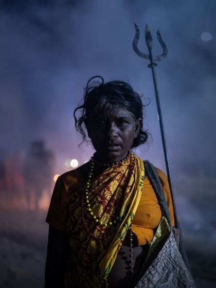 A woman holding a trishul stands amidst the evening smoke of a festival, her silhouette and direct gaze creating a powerful and mystical portrait.
