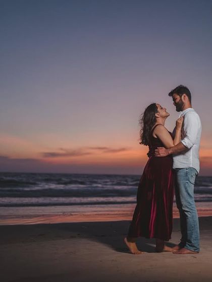 A full-length portrait of a couple on the beach at dusk. The deep colors of the sky and her velvet dress create a dramatic, cinematic feel.