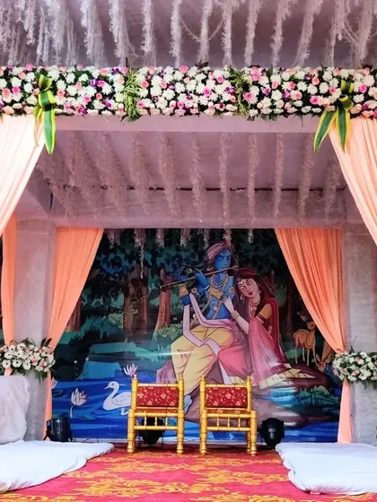 A unique vidhi mandap with a beautiful painted backdrop of Radha and Krishna. The stage is framed with peach-colored drapes and floral bouquets, adding a divine and artistic touch to the wedding ceremony.