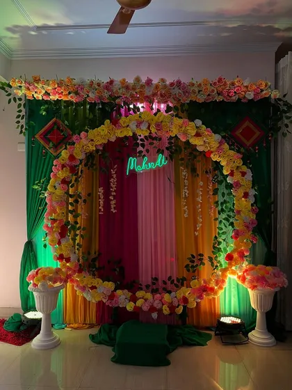 A vibrant and colorful Mehndi ceremony backdrop. This setup uses a floral ring, multi-colored drapes, and traditional elements to create a festive and photo-ready space for the bride-to-be.