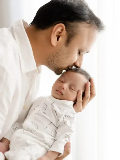 A classic father and newborn portrait. The father gently kisses his sleeping baby's forehead, a timeless expression of paternal love.