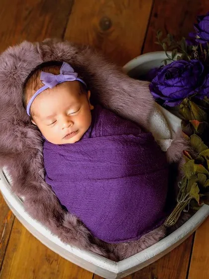 A sleeping baby swaddled in a rich purple wrap. The heart-shaped prop and matching flowers complete this beautifully styled shot.
