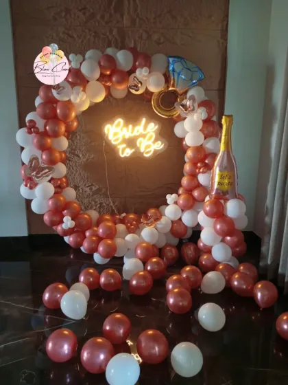 A chic 'Bride to Be' party decoration featuring a circular balloon arch in rose gold and white. The look is completed with a neon sign, a diamond ring foil balloon, and a champagne bottle accent.