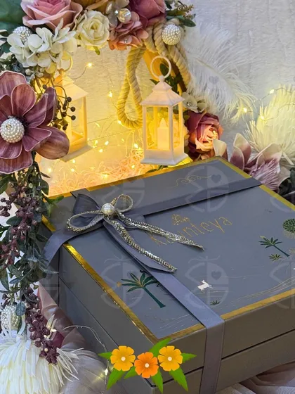 A beautifully packaged gift box for a wedding, featuring a custom grey box with a silver ribbon and delicate floral arrangements. The warm glow of miniature lanterns adds a magical touch to the presentation.