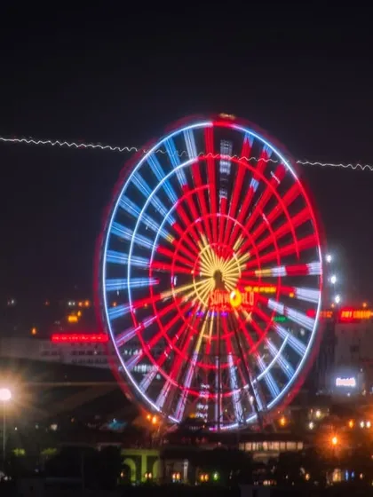 A long-exposure photograph of a brightly lit Ferris wheel in Vietnam, with the light trail of an airplane flying behind it.