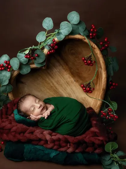 A beautiful composition with a baby wrapped in green, sleeping in a heart-shaped wooden bowl. The red berries and eucalyptus leaves create a lovely, natural frame.