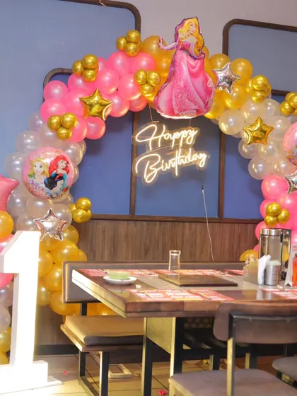 A princess-themed first birthday setup in a restaurant, featuring a pink and gold balloon arch, a princess foil balloon, and a 'Happy Birthday' neon sign.