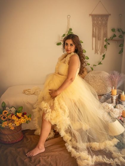 A full-length portrait showcasing the beautiful yellow tulle gown in the bohemian studio setting. The mix of textures, from the gown to the macrame and flowers, creates a visually rich and warm image.