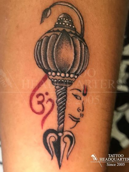 A composite spiritual tattoo featuring Hanuman's Gada (mace), a Trishul, the Om symbol, and a depiction of a deity's face.