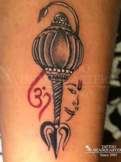 A composite spiritual tattoo featuring Hanuman's Gada (mace), a Trishul, the Om symbol, and a depiction of a deity's face.