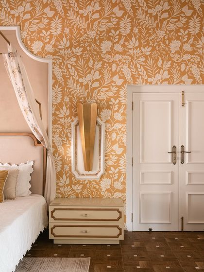 A closer look at the bedroom in Verdant Manor. The custom nightstand with gold trim and the sculptural brass wall sconce complement the bold floral wallpaper and the soft pink scalloped headboard.