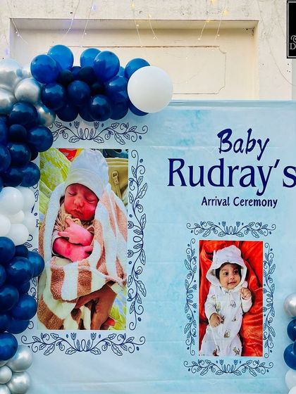 A personalized welcome backdrop for Baby Rudray's arrival ceremony. It features adorable baby photos framed by a blue and silver balloon garland, creating a heartfelt welcome.