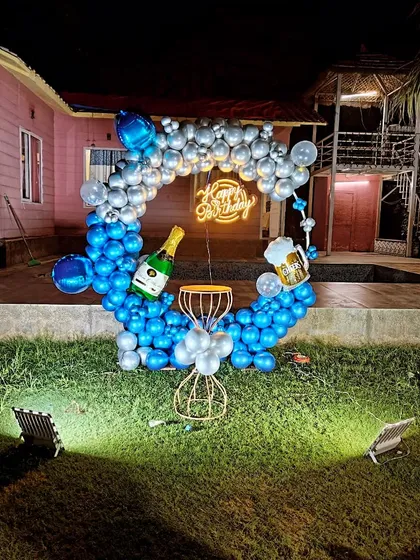 Here is an example of an outdoor birthday setup perfect for evening celebrations. This features a circular balloon frame in blue and silver, fun foil balloons like a champagne bottle, and a neon sign, all spotlit for a dramatic effect in a garden setting.
