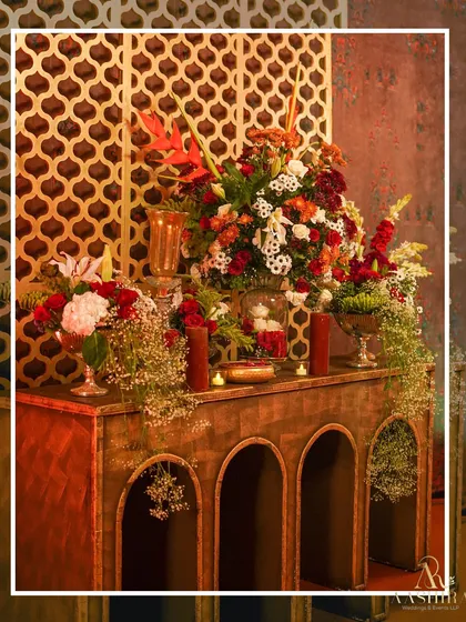 A beautiful console table with an arched design, laden with lush floral arrangements of red and white blooms, set against a golden jali screen.