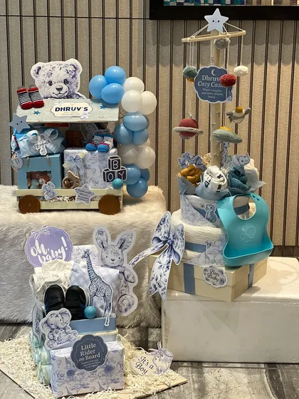 An overview of a custom baby arrival collection for a boy named Dhruv. It includes a trolley hamper, a diaper cake with a cozy mobile, and a mini wardrobe, all in a coordinated blue and white print.