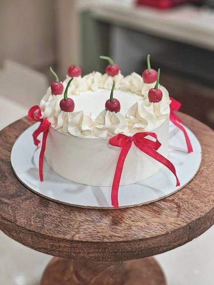 A whimsical cake with a cherry theme. It's topped with piped cream swirls, fondant cherries, and tied with little red ribbons for a fun and festive look.