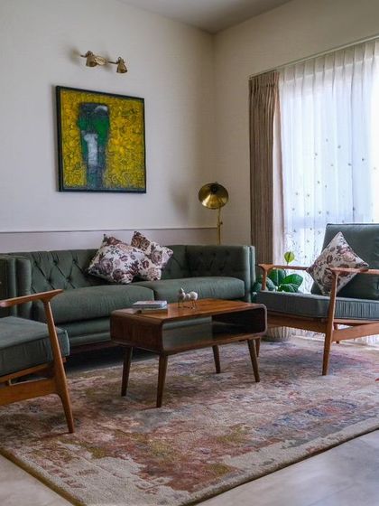 The living room of the 'Earthy Chic Bachelorette Pad,' where a Forest Green tufted sofa and traditional teak wood lounge chairs create a delightful fusion of warmth and earthy ambiance against a soft white paneled wall.