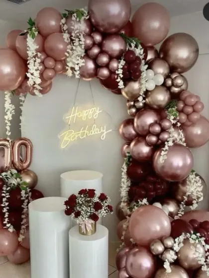 A luxurious 70th birthday celebration. The arch of rose gold and burgundy balloons is accented with white flowers and a 'Happy Birthday' neon sign for an elegant look.