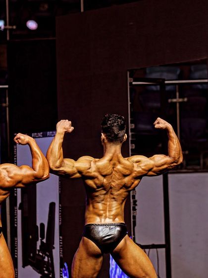 A back double biceps pose next to a competitor. This is a clear display of back width, density, and arm development, all key components of a bodybuilding contest.