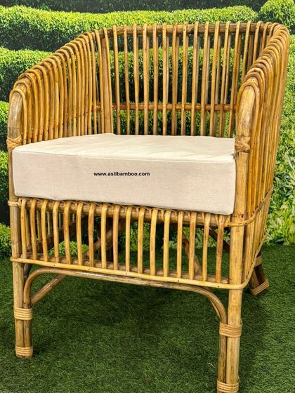 Our outdoor-ready rattan armchair with a simple cream cushion, set against a lush green backdrop. The natural bamboo frame is treated to be durable, making it a perfect choice for garden or patio seating.