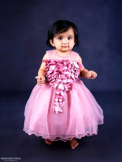 A baby girl stands proudly in her beautiful pink dress with floral details against a classic dark backdrop.