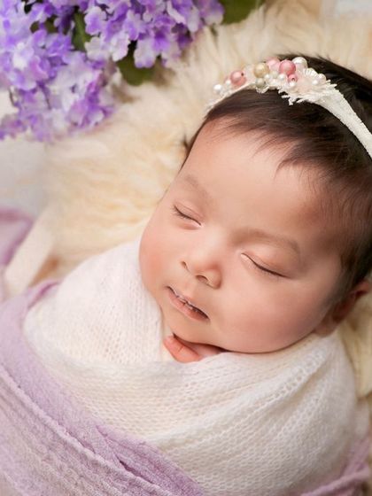 A close-up of a sleeping baby girl, adorned with a delicate pearl headband and surrounded by lavender flowers.