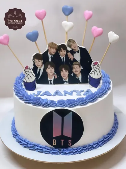 A BTS theme cake for a true fan. This cake is decorated with the BTS logo, a photo of the band, and heart-shaped lollipops, all on a whipped cream frosted cake.